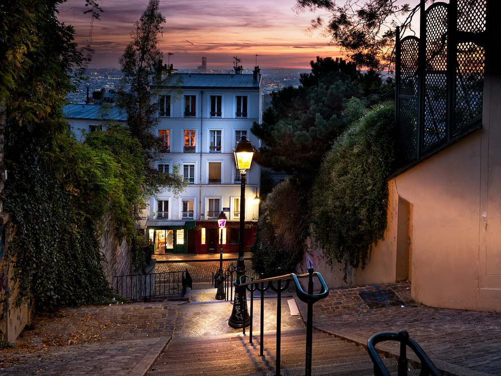 A picturesque Parisian street at sunset, featuring cobblestone steps, a quaint building, and a glowing street lamp, surrounded by lush greenery.