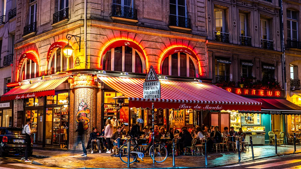 A vibrant Parisian caf&eacute;, Bar du March&eacute;, illuminated at night with red and white striped awnings and outdoor seating filled with patrons enjoying the lively atmosphere.