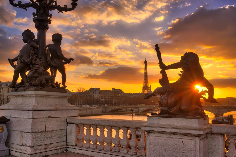 A stunning sunset over the Seine River in Paris, featuring intricate sculptures and the Eiffel Tower silhouetted against a colorful sky.