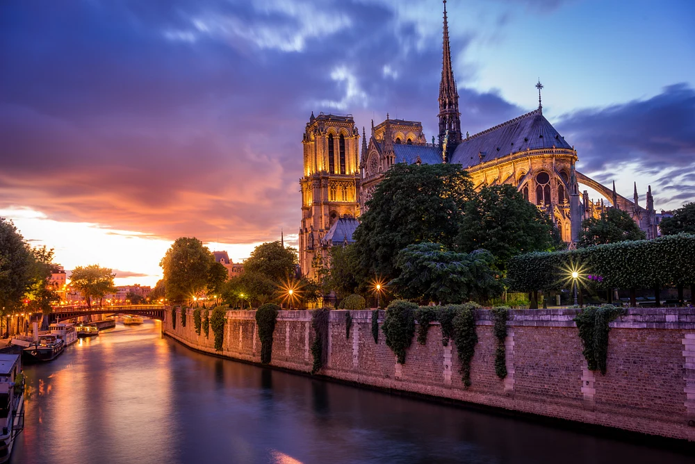 A stunning view of Notre-Dame Cathedral at twilight, illuminated by golden light, with the Seine River reflecting the vibrant sunset hues and framed by lush trees.
