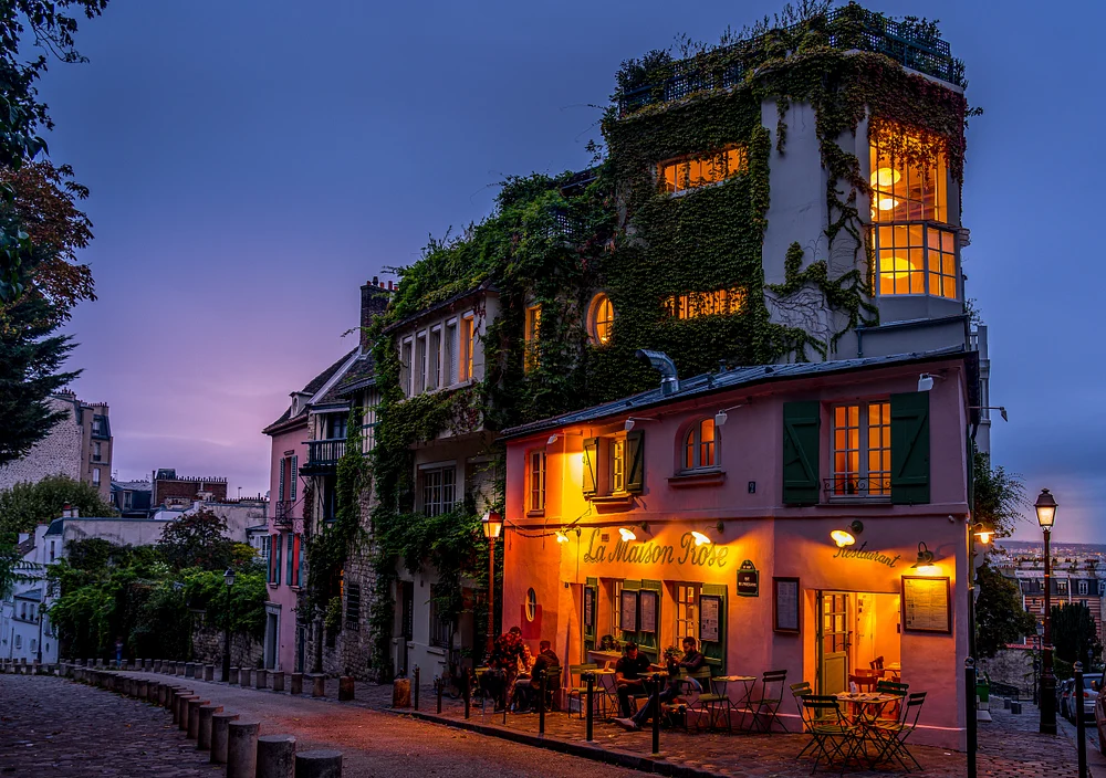 A beautifully illuminated Parisian caf&eacute; at dusk, adorned with greenery and inviting outdoor seating, set against a soft evening sky.