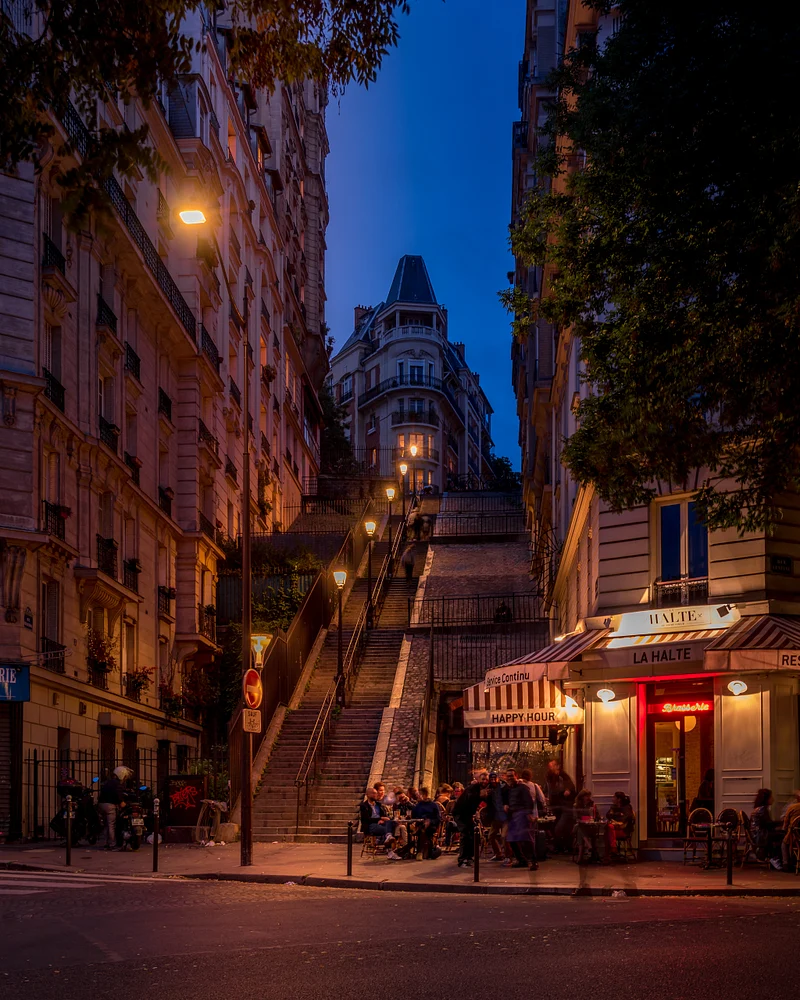 A bustling street corner in Montmartre, Paris, featuring a grand staircase illuminated by streetlights, with patrons enjoying a caf&eacute; in the foreground during twilight.