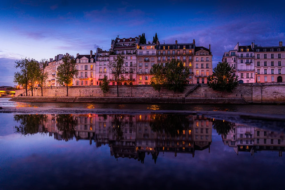 A serene view of the Parisian riverfront at dusk, showcasing elegant buildings reflecting in the calm waters of the Seine, framed by trees and illuminated by soft evening lights.