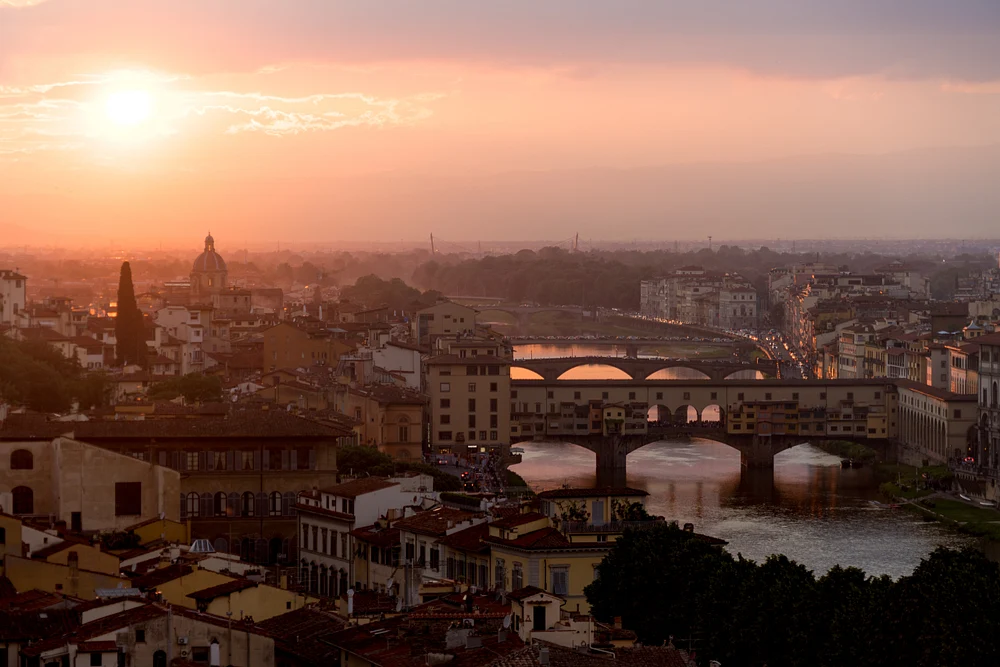 A stunning sunset over Florence, showcasing the Arno River and the Ponte Vecchio bridge, with warm hues reflecting off the water and historic rooftops in the foreground.
