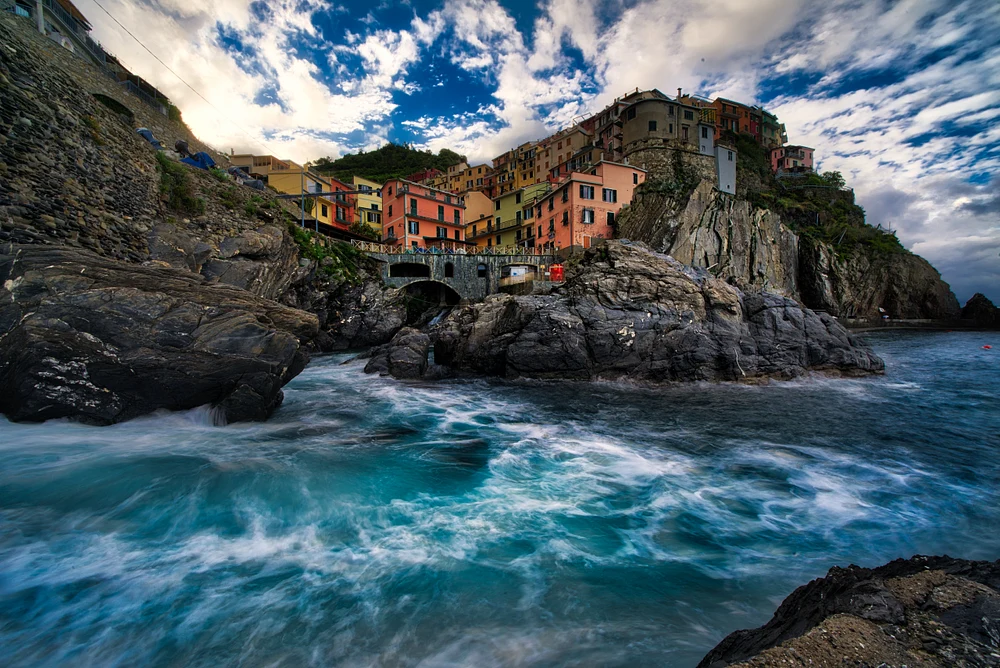 A stunning view of Manarola, a coastal village in Cinque Terre, showcasing colorful houses on cliffs, crashing waves, and a dramatic sky.