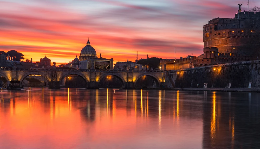 A stunning sunset over the Tiber River in Rome, featuring St. Peter's Basilica and Castel Sant'Angelo, with vibrant colors reflecting on the water.
