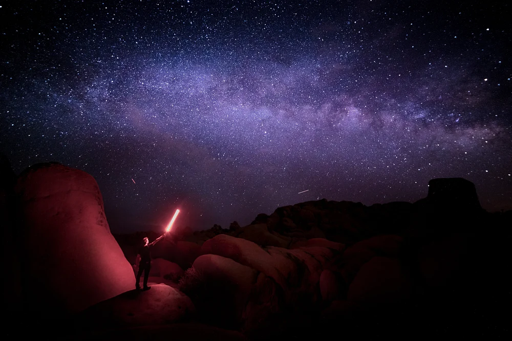 A lone Jedi stands in a desert at night, holding a glowing lightsaber under a starry sky filled with the Milky Way, surrounded by rugged rocks.