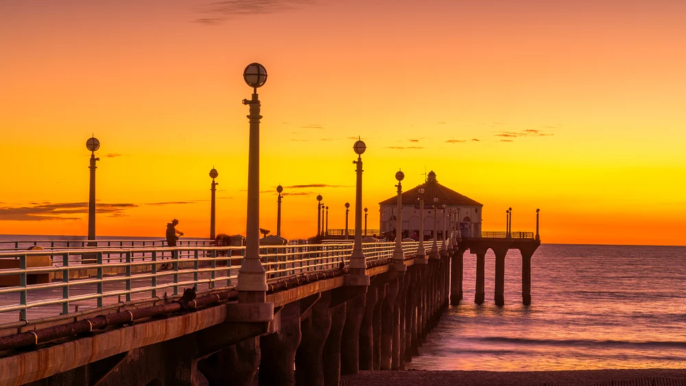 A golden sunset over Manhattan Beach Pier, featuring silhouetted lampposts and a pavilion against a vibrant orange and yellow sky.