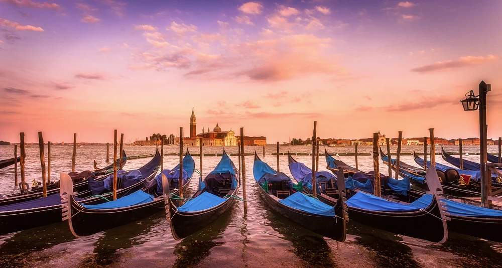 A fleet of blue gondolas moored along wooden poles in Venice at twilight, with San Giorgio Maggiore church in the background under a colorful sunset sky.