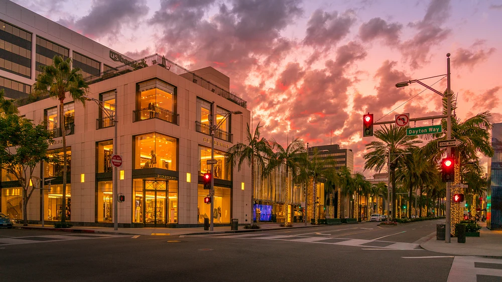 A stunning view of modern retail architecture on Dayton Way in Beverly Hills during golden hour, featuring palm trees and a colorful sunset sky.