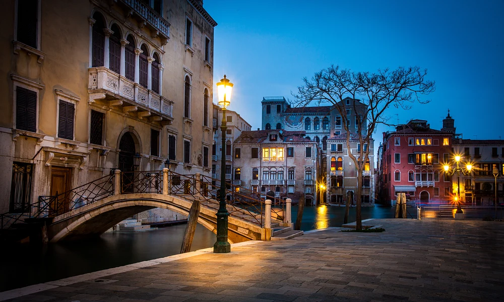 A beautifully lit bridge over a tranquil canal in Venice at dusk, surrounded by historic buildings and glowing street lamps.