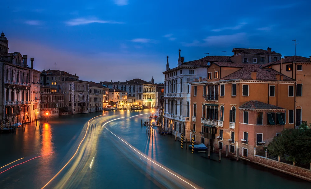 A tranquil evening scene in Venice, showcasing the illuminated canals and historic buildings under a soft blue sky, with trails of boat lights reflecting on the water.