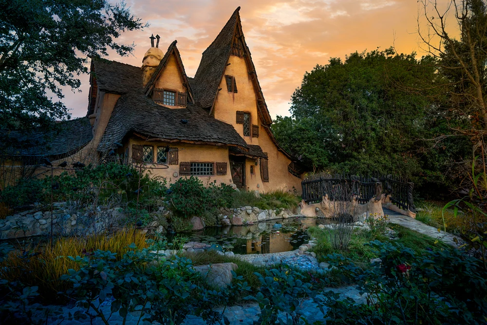 A whimsical cottage with pointed roofs surrounded by greenery and a pond at sunset, creating a magical atmosphere.