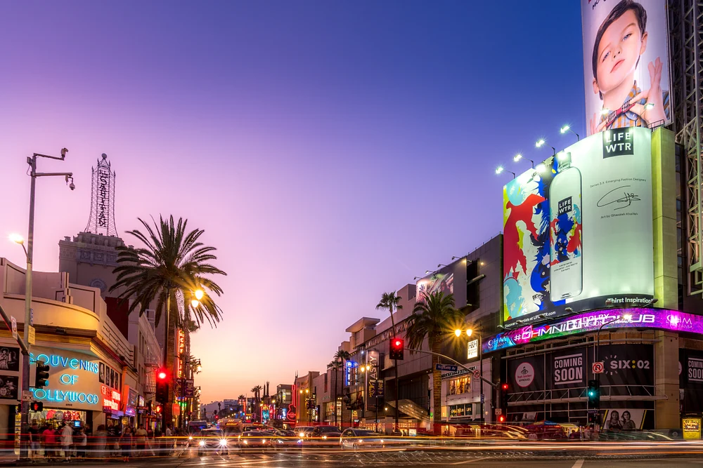 A vibrant sunset over Hollywood Boulevard, showcasing palm trees, neon signs, and bustling traffic, capturing the lively essence of Los Angeles' entertainment hub.