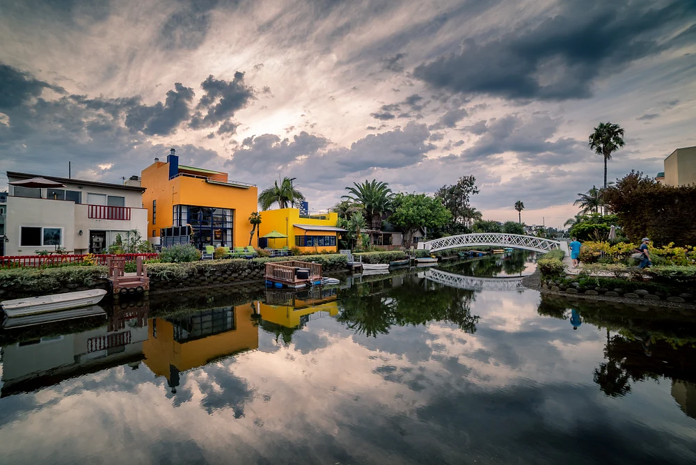 A picturesque view of the Venice Canals in Los Angeles at sunset, featuring vibrant yellow buildings reflecting in the calm water, framed by a white arched bridge and palm trees under a dramatic cloudy sky.