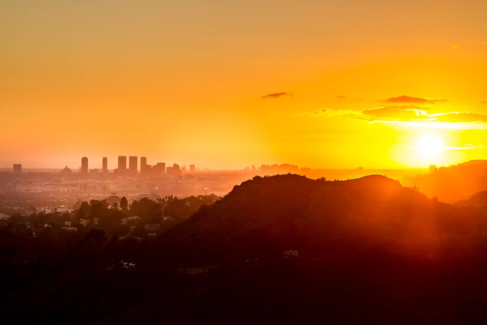 A stunning sunset over the Los Angeles skyline, featuring vibrant orange and yellow hues with rolling hills in the foreground.