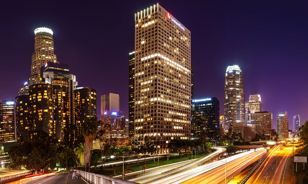 Nighttime view of the Los Angeles skyline with light trails from vehicles, showcasing iconic skyscrapers and palm trees against a purple sky.