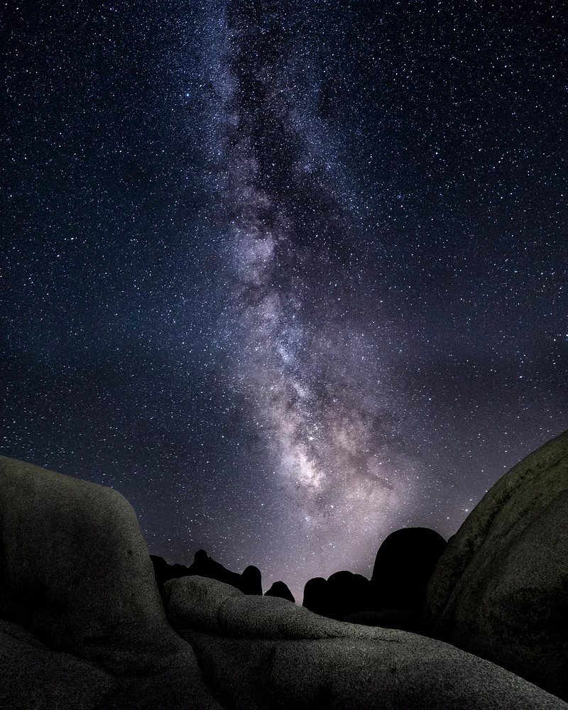A stunning view of the Milky Way galaxy arching over ancient desert rocks, filled with countless stars against a deep indigo and lavender sky.