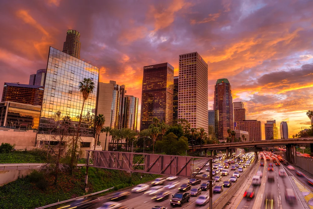A stunning sunset over Downtown Los Angeles, featuring vibrant orange and purple hues illuminating the skyline and a busy freeway below with streaks of headlights and taillights.