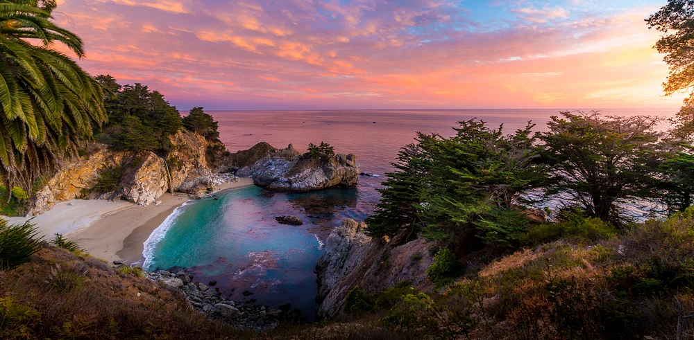 A stunning sunset over McWay Cove in Big Sur, showcasing turquoise waters, dramatic cliffs, and vibrant sky colors.