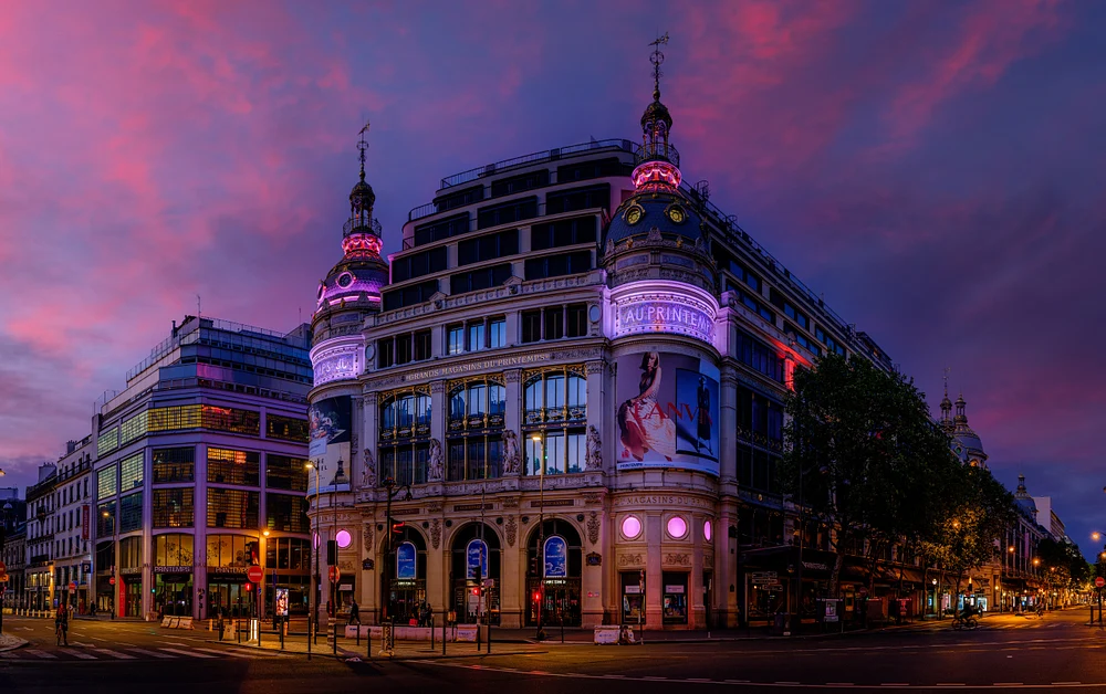 Evening view of Le Printemps department store in Paris, showcasing its illuminated fa&ccedil;ade against a colorful sunset sky.