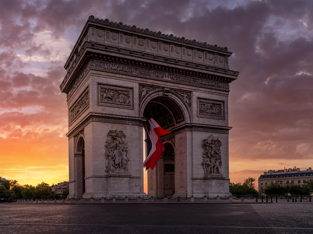 The Arc de Triomphe in Paris illuminated by sunset, featuring the French flag and a vibrant sky.