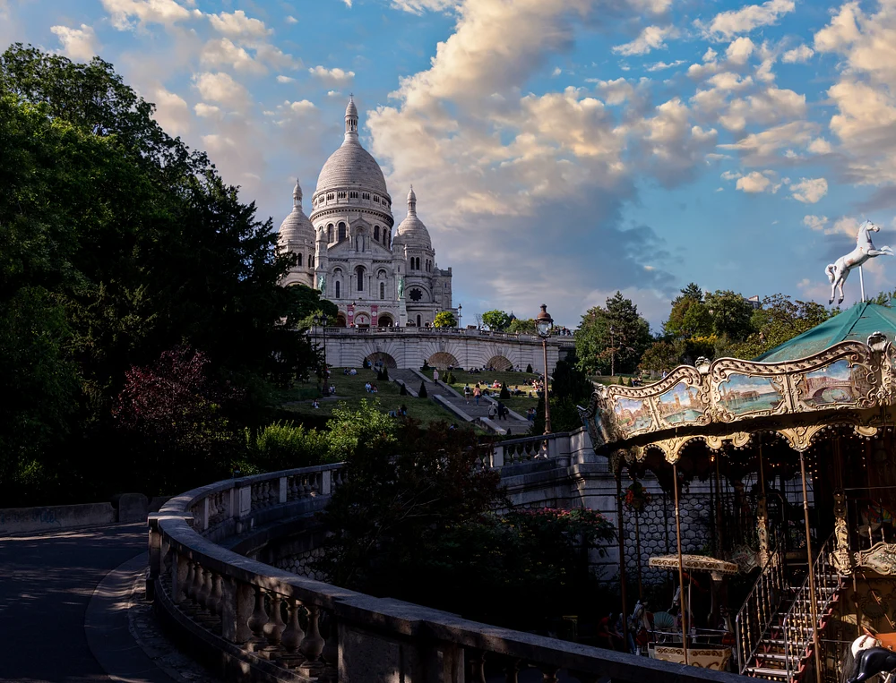 Scenic view of Sacr&eacute;-C&oelig;ur Basilica with a carousel in Montmartre, Paris, showcasing lush greenery and a beautiful sky.