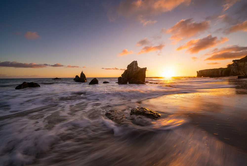 A serene beach sunset featuring rocky outcrops, gentle waves, and a colorful sky transitioning from gold to pink and blue.