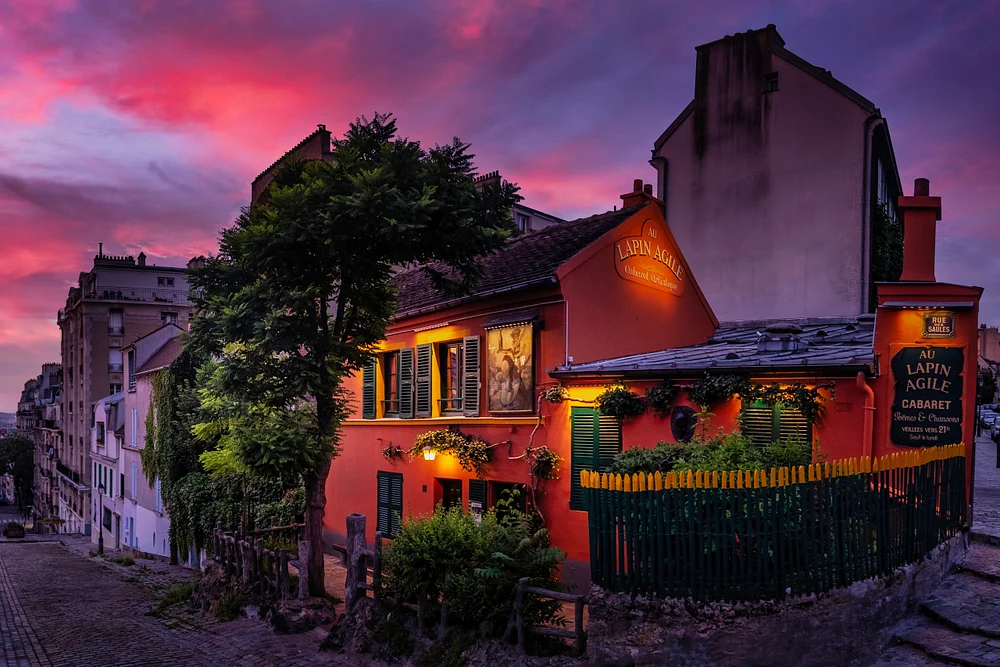 A picturesque view of Lapin Agile cabaret in Montmartre, Paris, illuminated at dusk with a stunning pink and purple sky in the background.