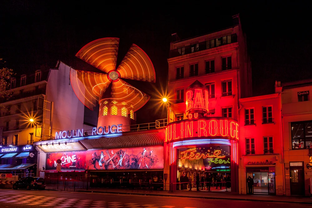 Night view of the Moulin Rouge in Paris, showcasing its illuminated red facade and iconic windmill against a dark sky.