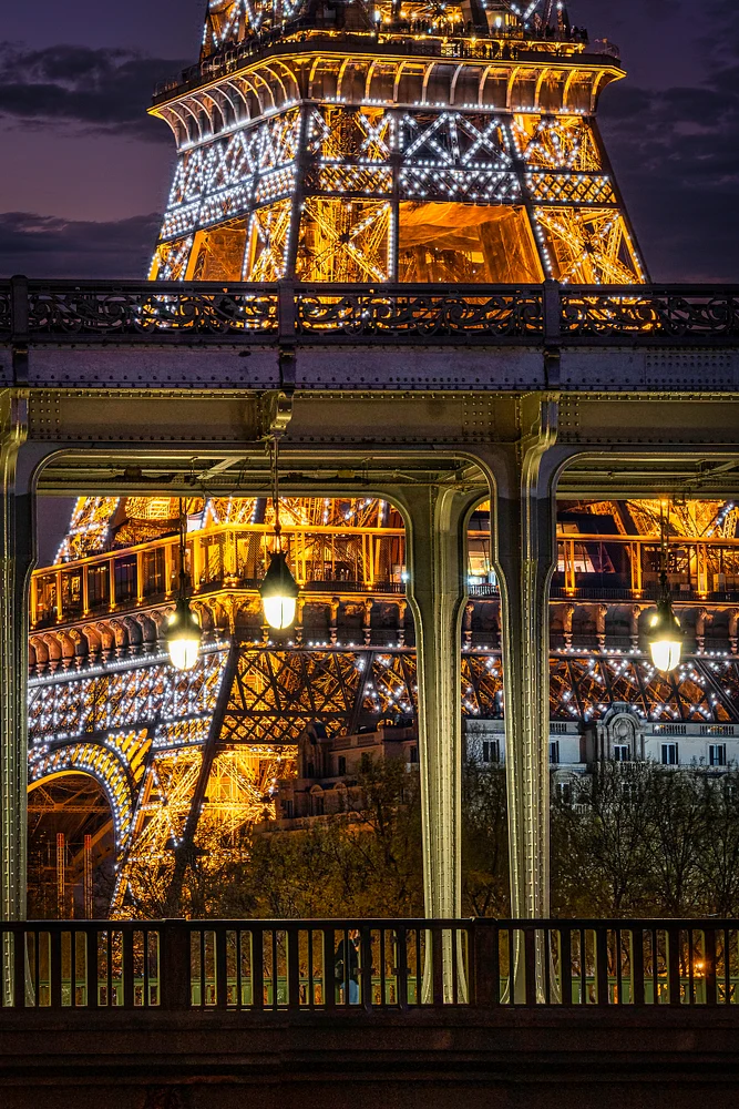 A close-up view of the Eiffel Tower illuminated at dusk, framed by a nearby bridge with glowing lanterns, showcasing the romantic charm of Paris.