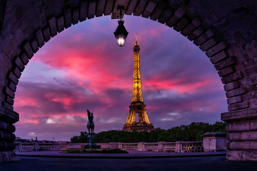 A stunning view of the Eiffel Tower at sunset, framed by a stone archway, with a statue in the foreground and vibrant clouds in the sky.