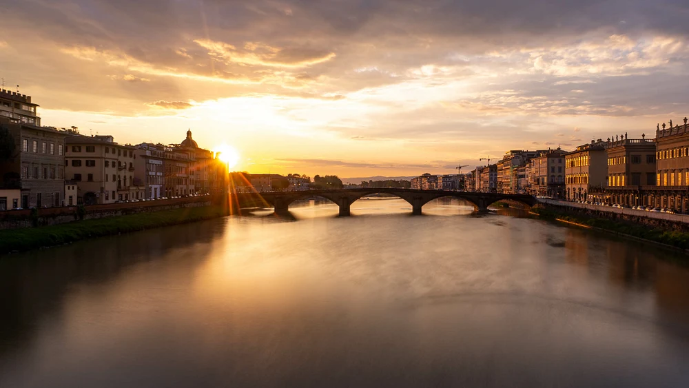 A stunning sunset over the Arno River in Florence, showcasing golden hues reflecting on the water with historic architecture lining the riverbanks.
