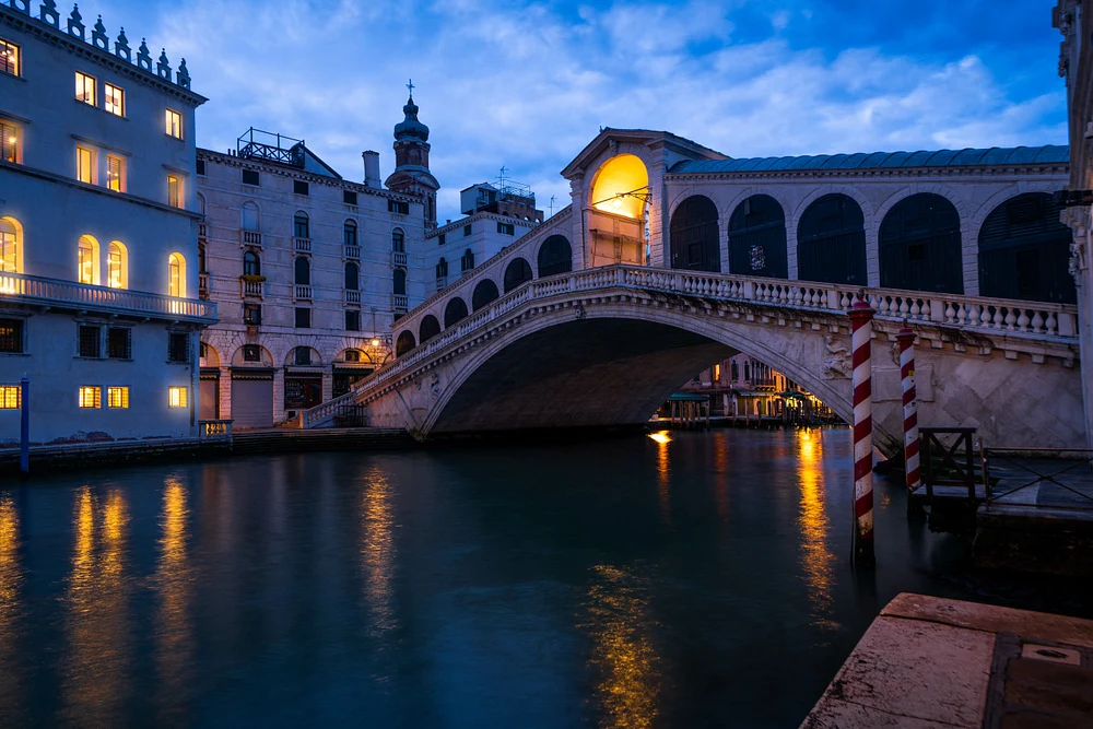 A twilight view of the Rialto Bridge in Venice, with reflections in the Grand Canal and illuminated buildings nearby.