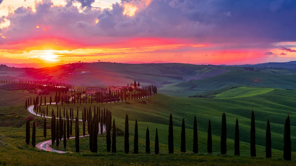 A stunning sunset over Tuscany's rolling hills, featuring a winding road lined with cypress trees and a charming villa amidst lush green fields.