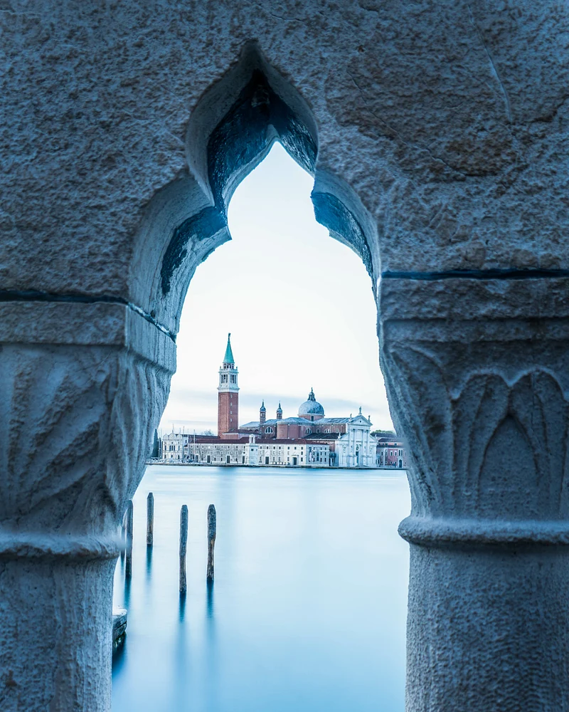 A serene view of Venice framed by an ornate archway, showcasing St. Mark's Basilica and calm waters in soft blue tones.