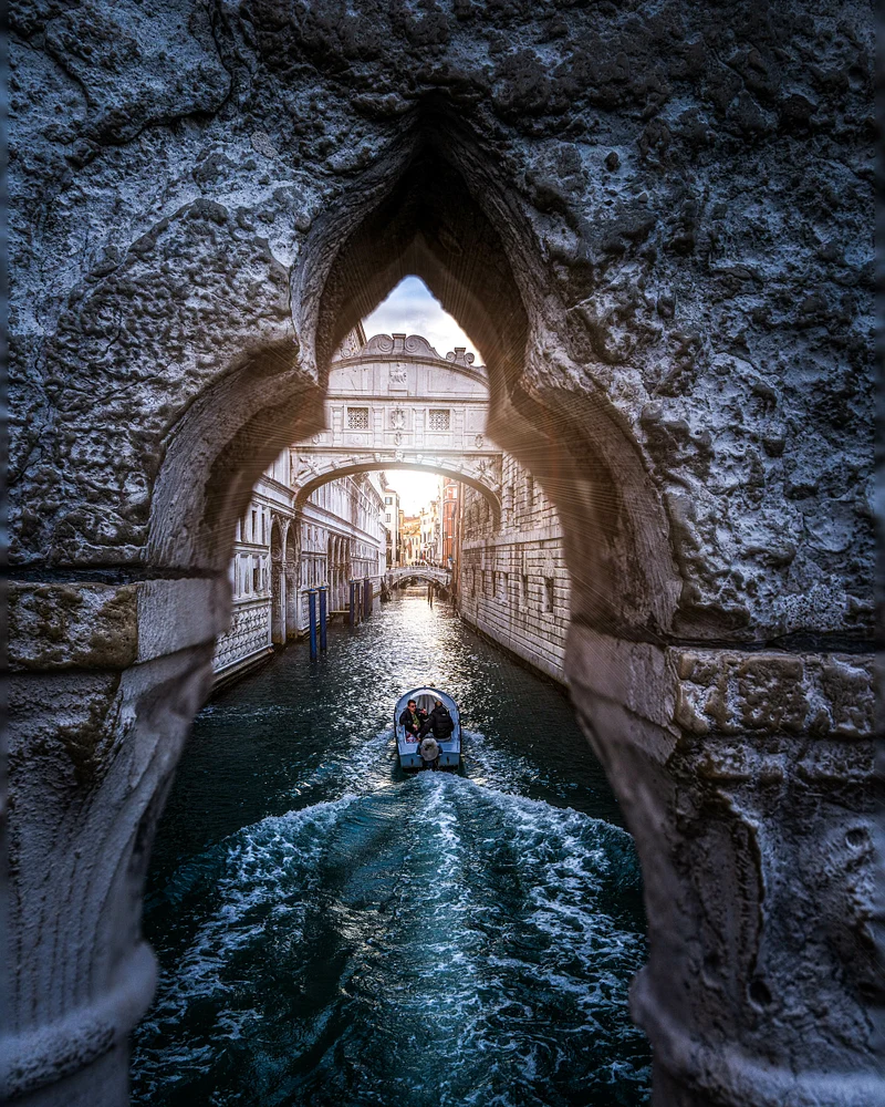 A view of the Bridge of Sighs in Venice framed by ancient stone, showcasing a serene canal at sunset with a boat gliding through the water.