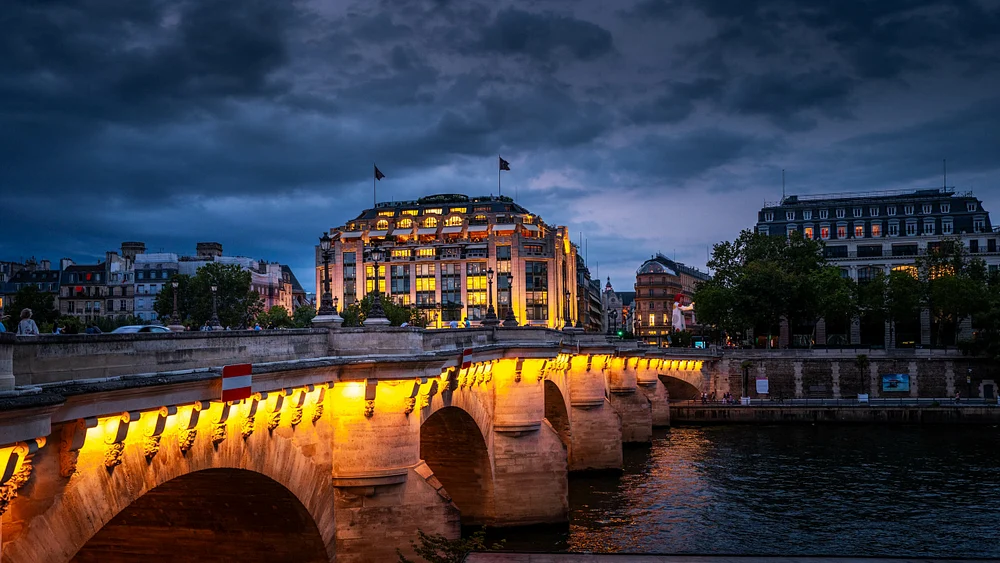 A stunning view of Pont Neuf illuminated at dusk, with La Samaritaine in the background, reflecting in the Seine River under a dramatic sky.