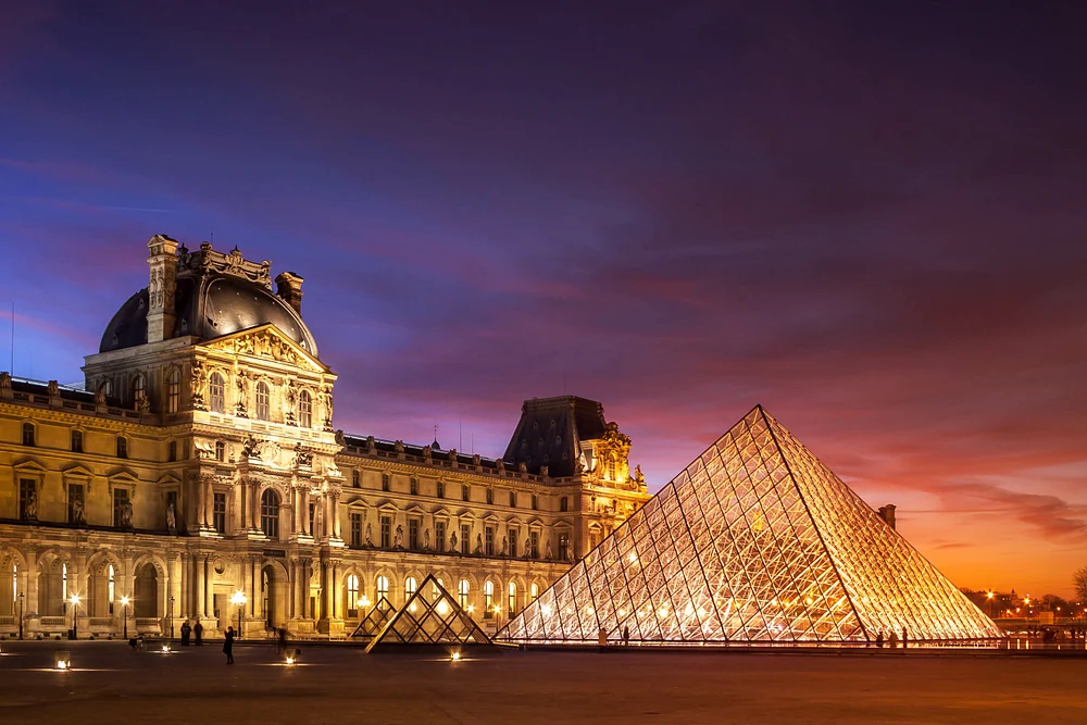 Sunset view of the Louvre Museum in Paris, featuring the iconic glass pyramid illuminated against a colorful sky.