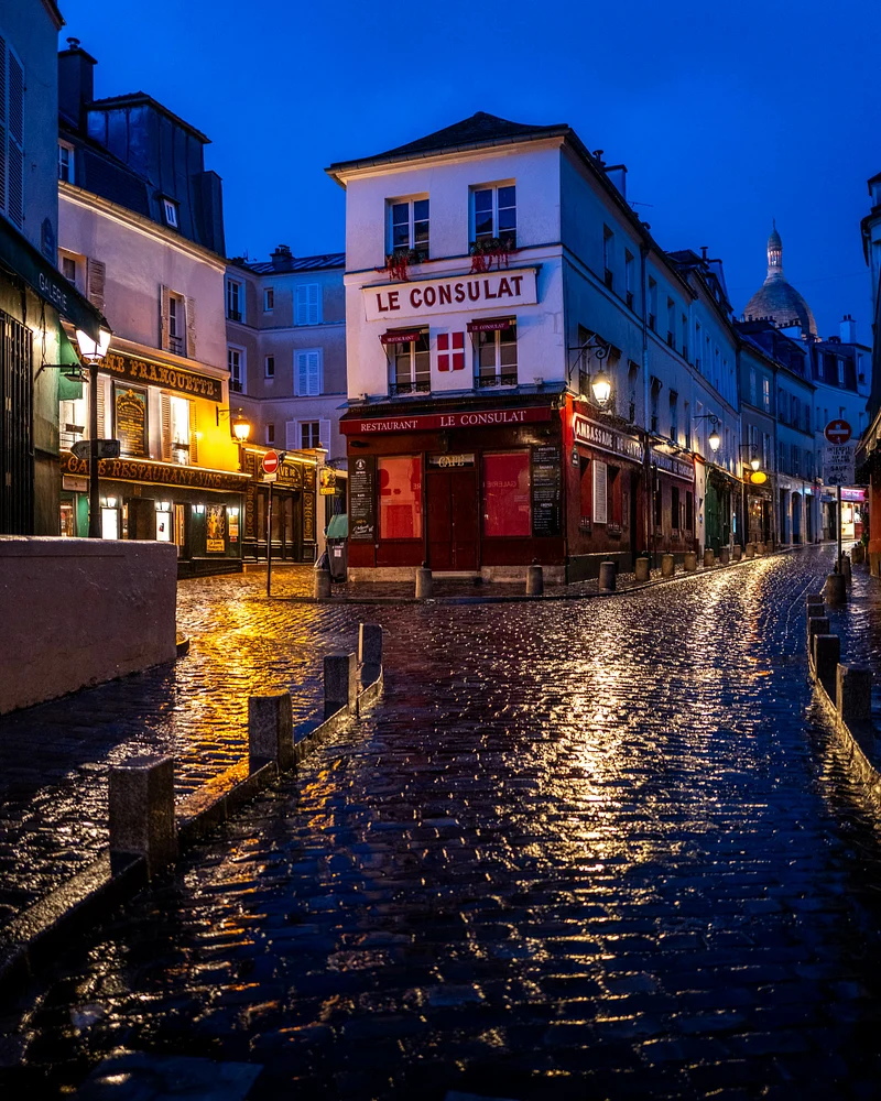Evening photograph of Montmartre, Paris, showcasing cobblestone streets glistening after rain, with the iconic restaurant