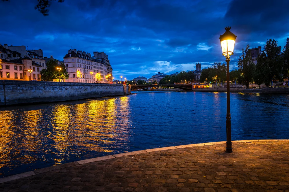 A serene evening view of the Seine River in Paris, showcasing shimmering reflections of city lights on the water and a glowing streetlamp along the riverbank.