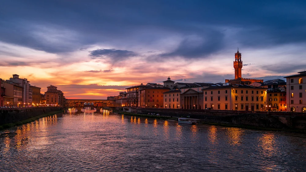 A stunning nighttime view of the Arno River in Florence, showcasing illuminated historic buildings and the silhouette of Palazzo Vecchio against a colorful sunset sky.