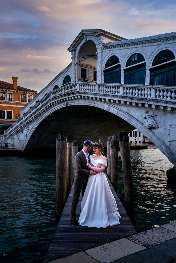 A couple shares a romantic moment on a wooden dock at the Rialto Bridge in Venice, with the bride in an elegant off-the-shoulder gown and the historic architecture in the background.