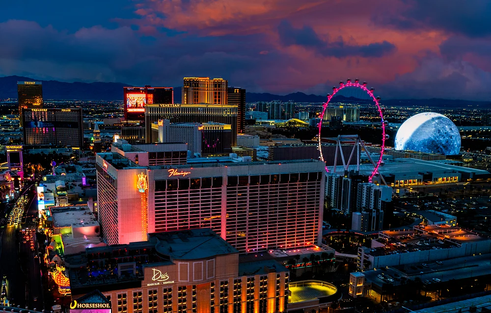 Aerial view of Las Vegas at dusk showcasing the Strip, High Roller Ferris wheel, and the Sphere at The Venetian against a colorful sunset backdrop.