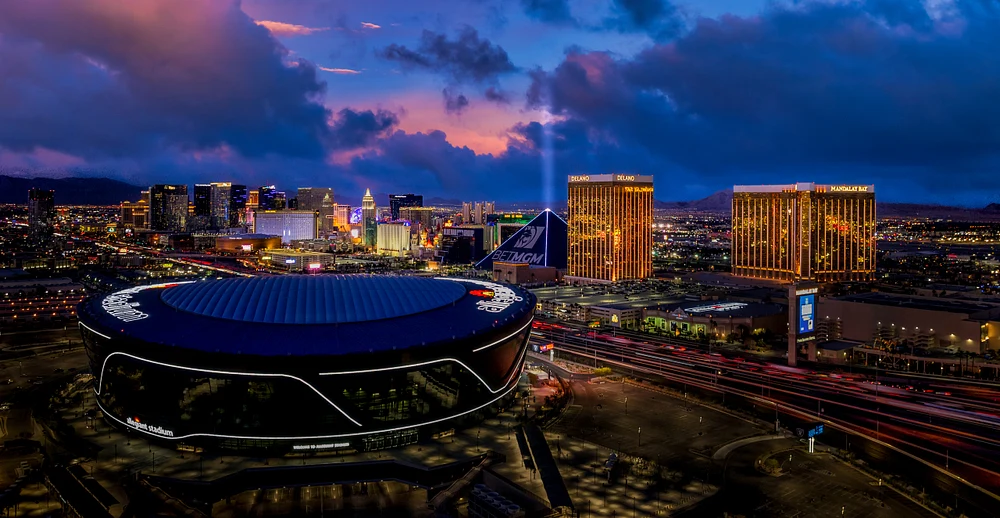 Aerial view of the Las Vegas skyline at dusk, featuring Allegiant Stadium and iconic buildings like the Luxor and MGM Grand, with colorful clouds in the background.