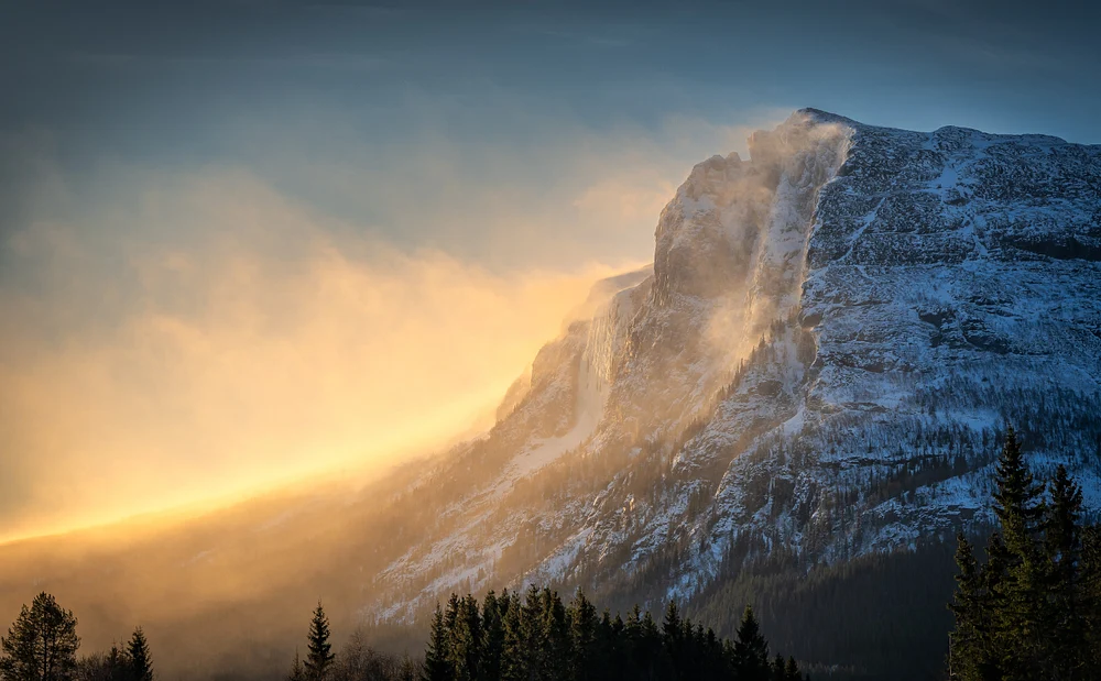 A stunning view of Vesleh&oslash;dn Mountain with snow drifting in the air, illuminated by a golden sunset, showcasing the beauty of winter landscapes.
