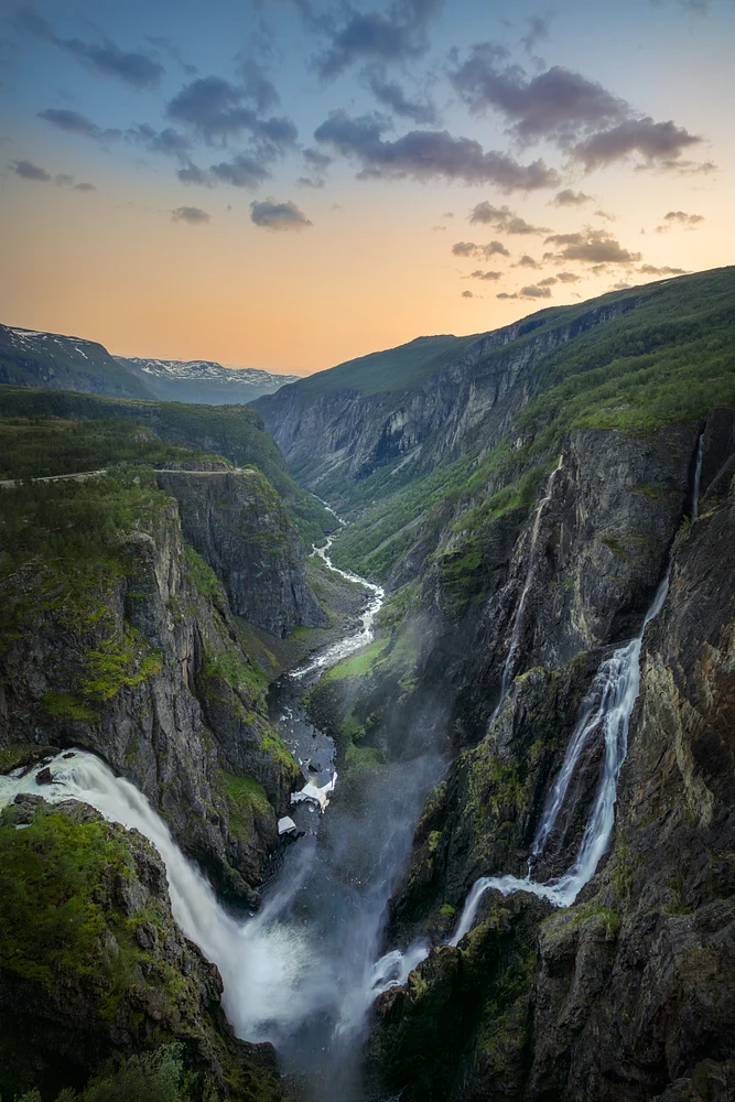 A stunning view of V&oslash;ringsfossen waterfall cascading into a lush valley at sunset, surrounded by rocky cliffs and greenery.