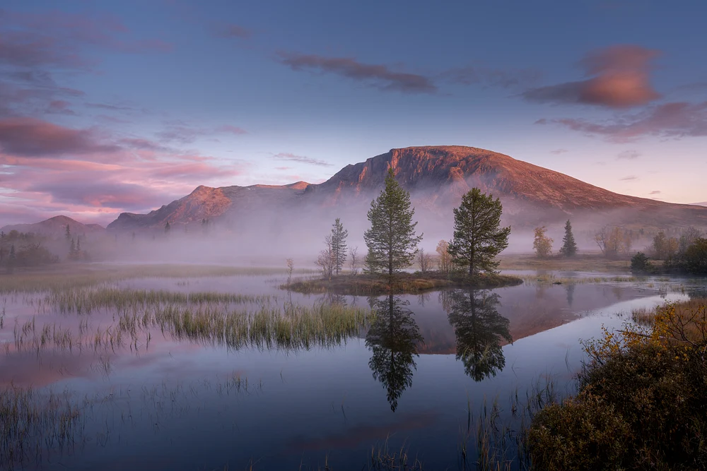A majestic mountain at sunrise, reflecting in tranquil waters with mist and trees surrounding the scene.
