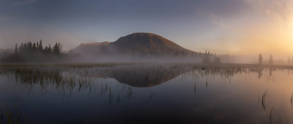 A panoramic view of Skogshorn Mountain shrouded in morning mist, reflecting in a calm lake surrounded by trees.