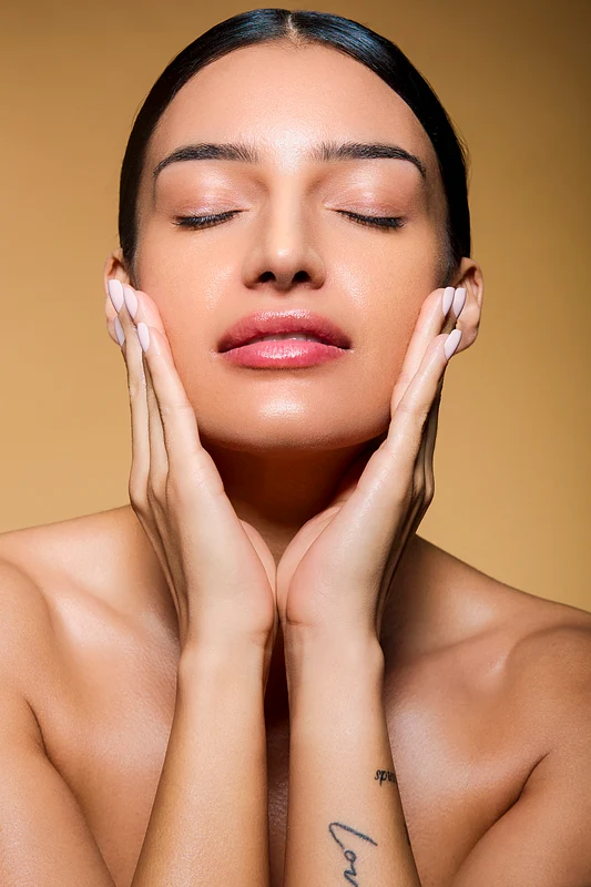 Glowing Model Embracing Natural Beauty in Studio A serene model with closed eyes gently holds her face, showcasing flawless skin against a warm background.
